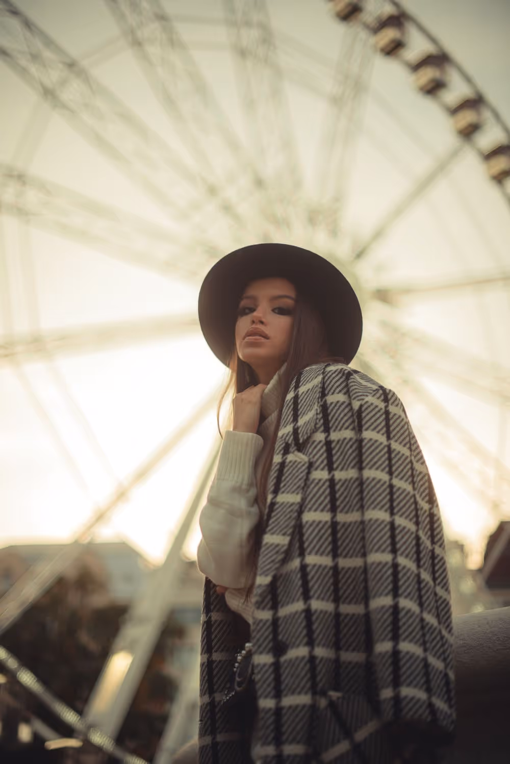Woman wearing a black hat and checkered coat posing outdoors with a Ferris wheel in the background at sunset.
