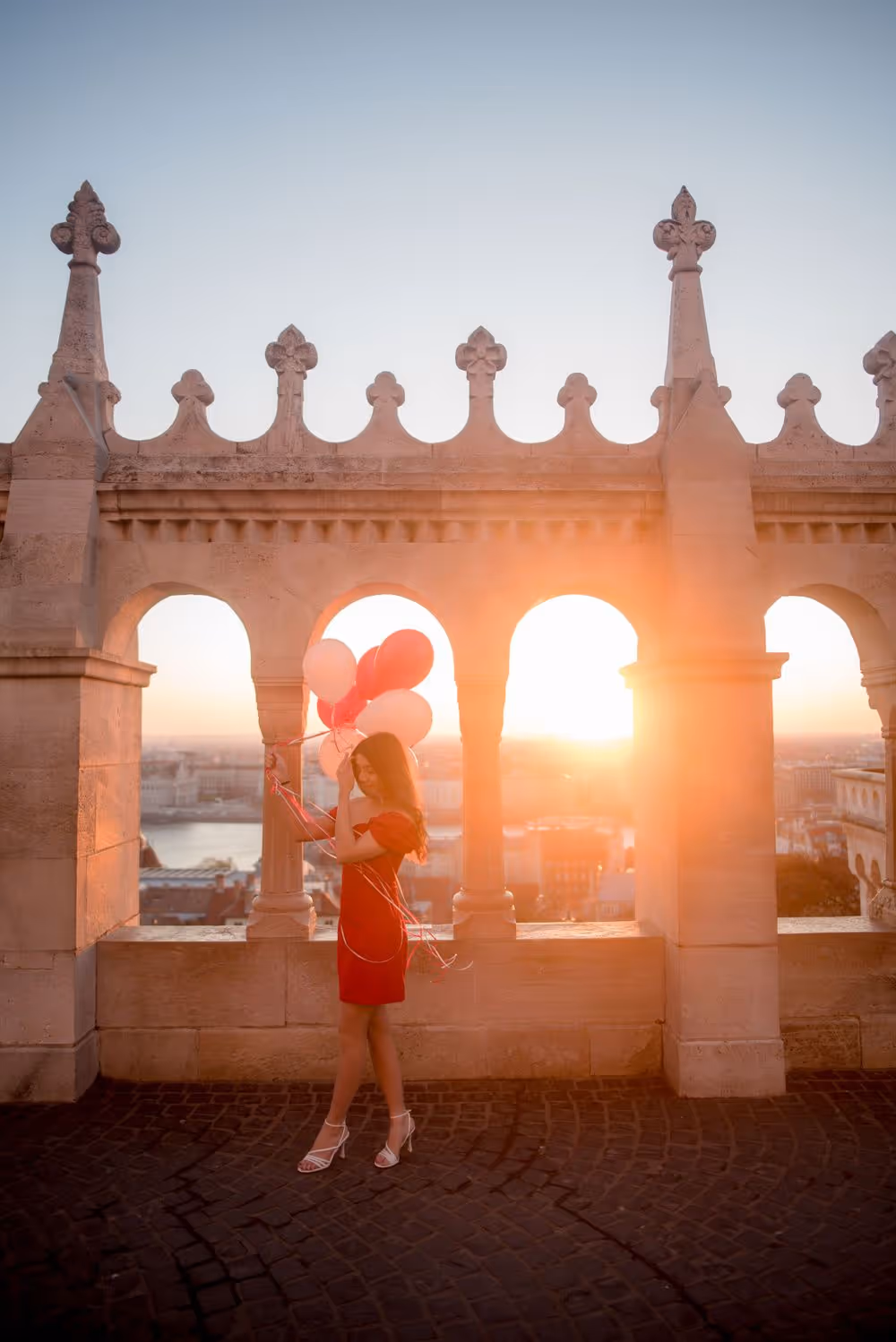 Woman in a red dress holding red and white balloons standing under decorative stone arches at sunset.