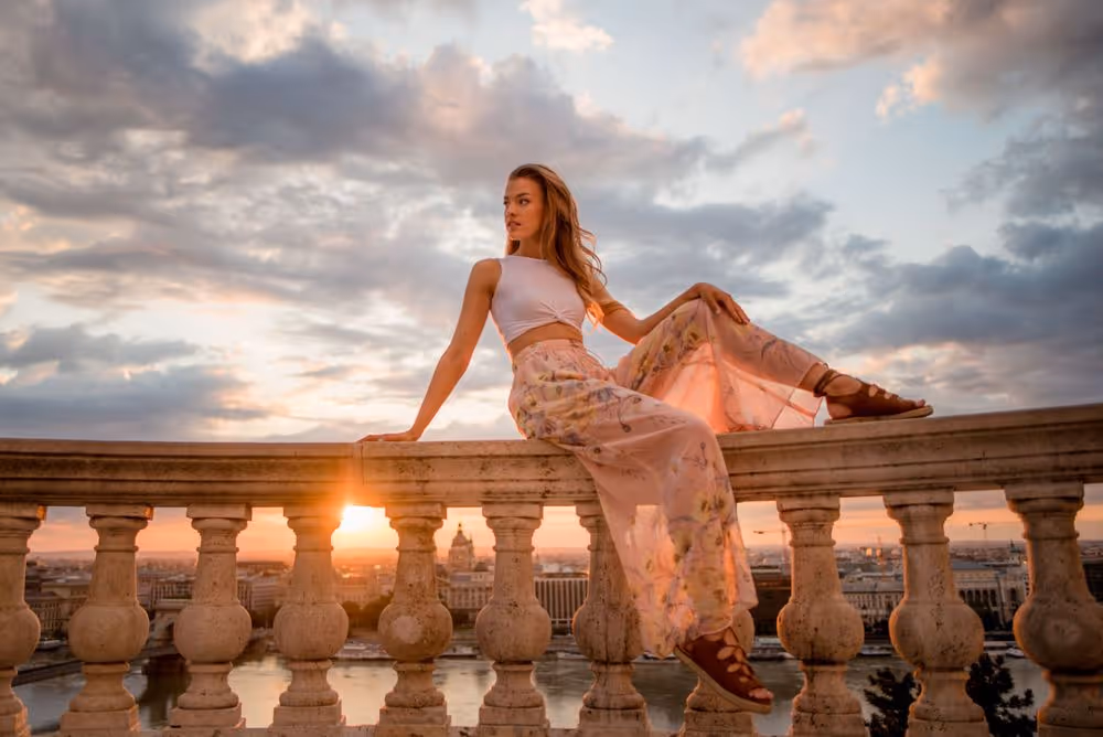 Young woman in a white crop top and floral pants sitting on a stone balustrade at sunset with clouds and cityscape in the background.