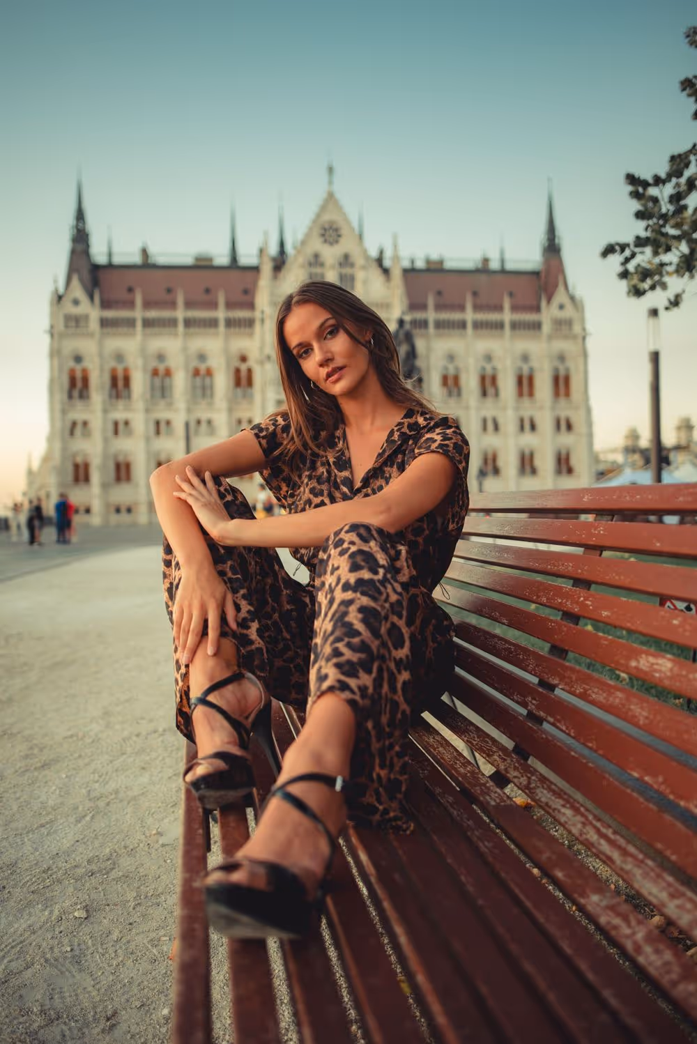 Woman wearing a leopard print outfit sitting on a wooden bench with a historic Gothic-style building in the background.