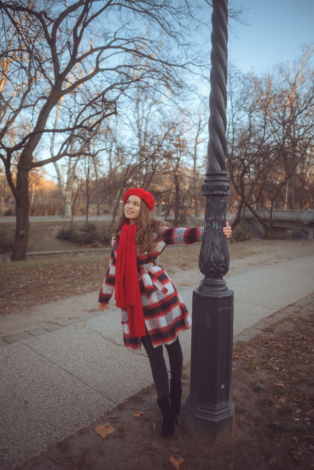 Young woman in red beret, scarf, and checkered coat holding a tall street lamp in a park during late autumn.