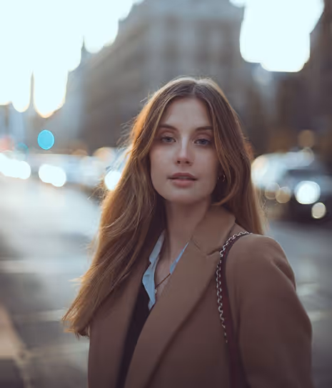 Young woman with long brown hair wearing a camel coat and black top standing outdoors on a city street at dusk.