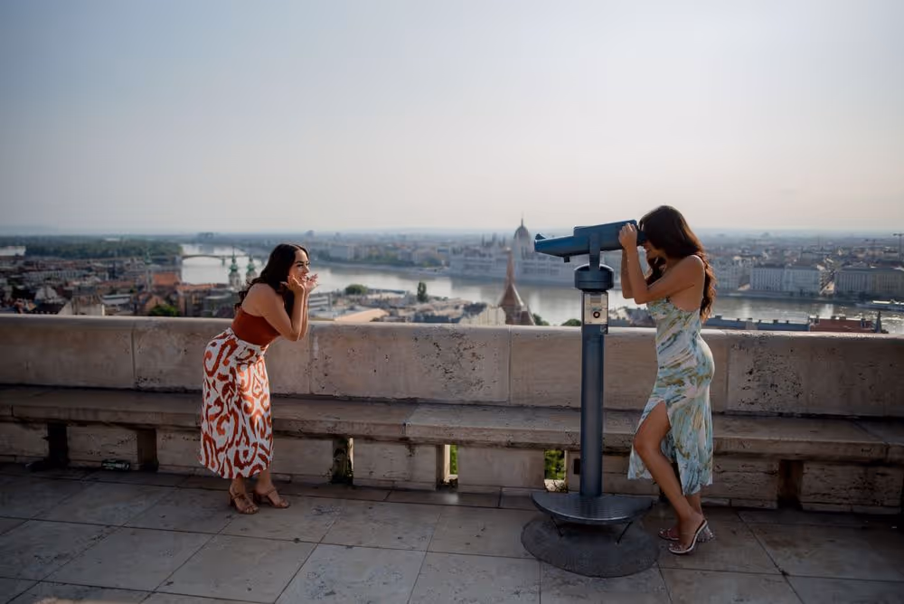 Two women on a terrace overlooking a river and cityscape, one looking through a public binocular viewer and the other posing playfully.