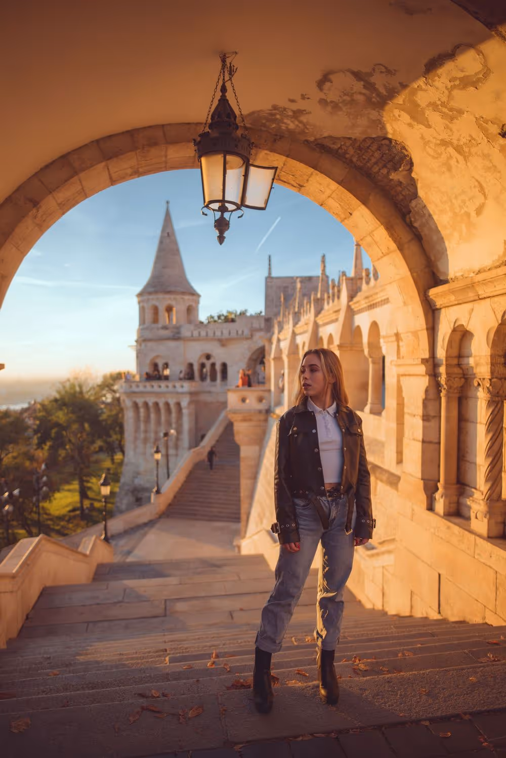 Woman in a black jacket and jeans standing on stone steps under an arched corridor at sunset with a historic castle tower in the background.