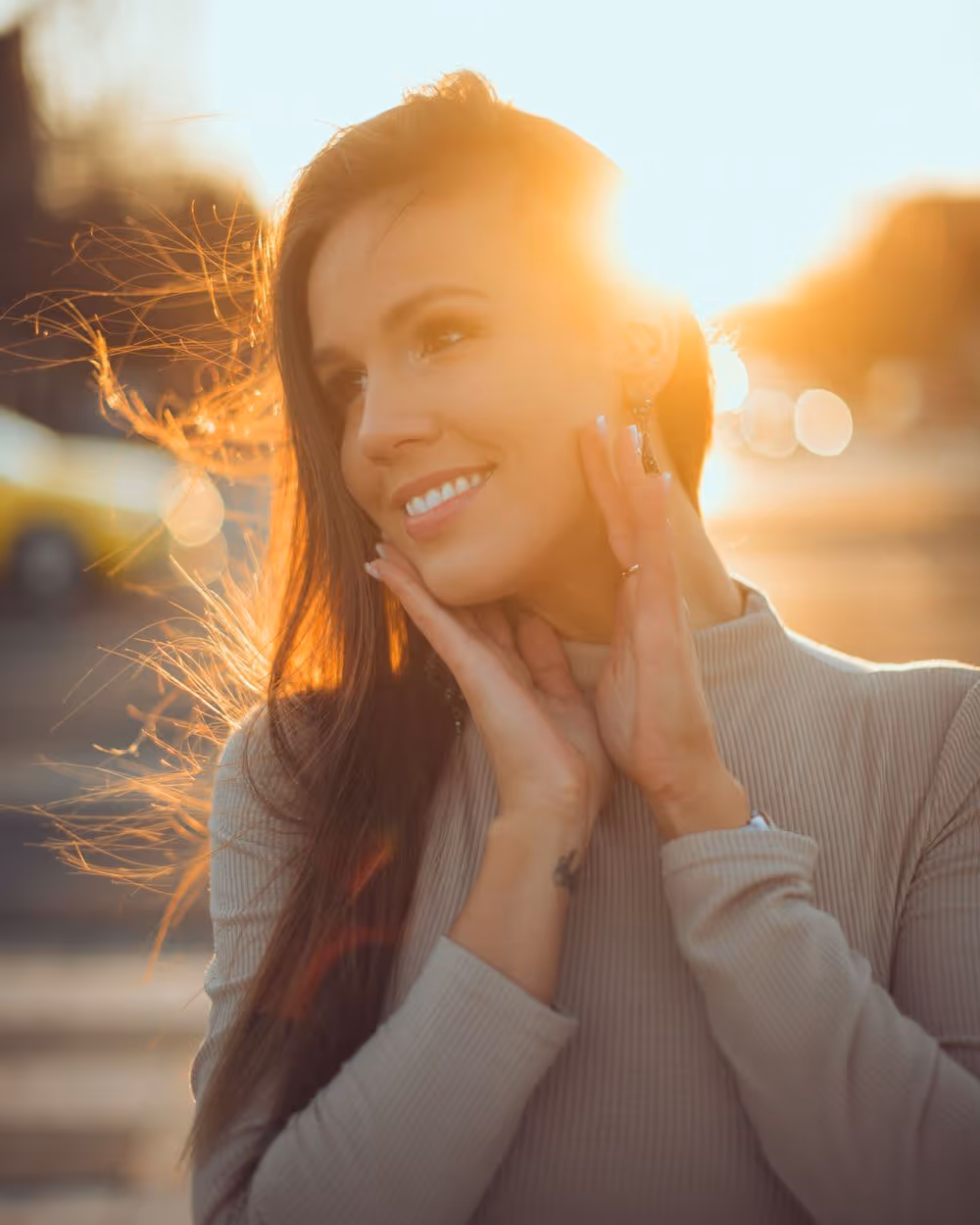 Smiling woman with long hair holding her face, backlit by warm sunlight creating a glowing effect.