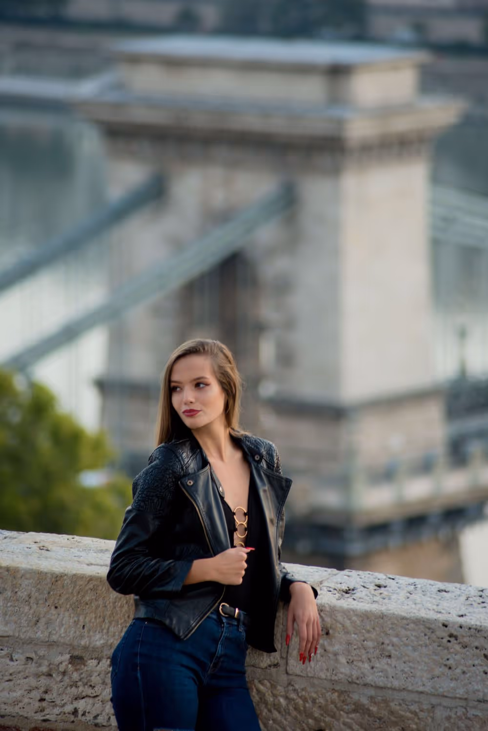 Young woman wearing a black leather jacket and jeans leaning on a stone railing with a blurred historic suspension bridge in the background.