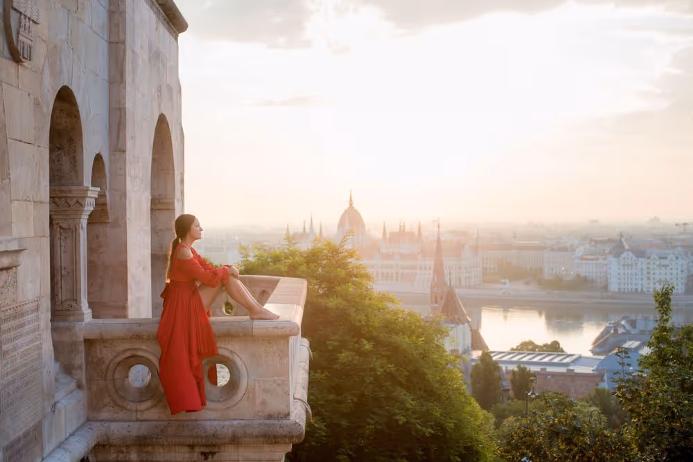A woman in a red dress sitting barefoot on a stone balcony overlooking a cityscape at sunrise or sunset.
