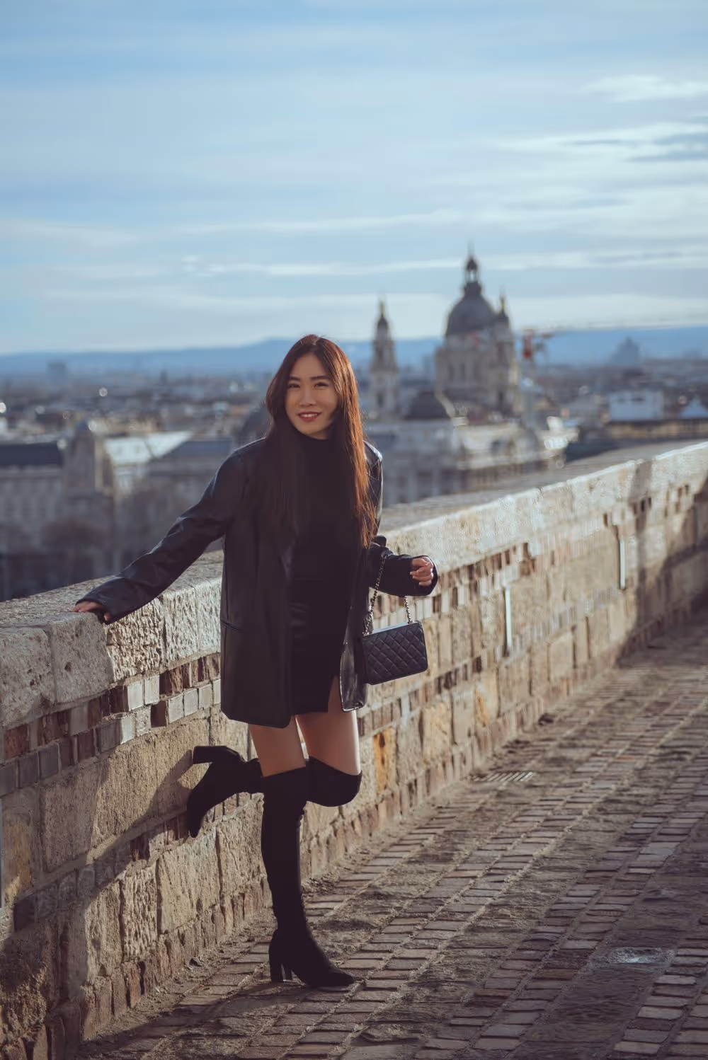 Woman in black leather jacket, short dress, and thigh-high boots posing on a stone walkway with a cityscape and historic buildings in the background.