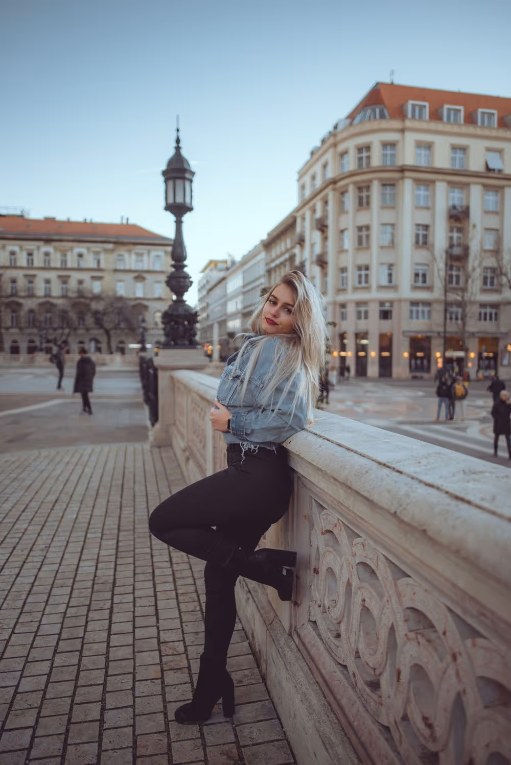 Young woman with long blonde hair leaning against ornate stone railing in urban setting with historic buildings.