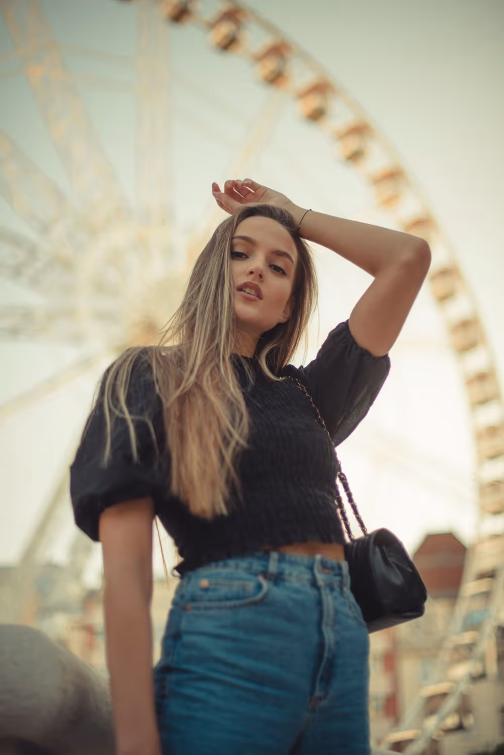 Young woman in a black top and blue jeans posing with a Ferris wheel blurred in the background.