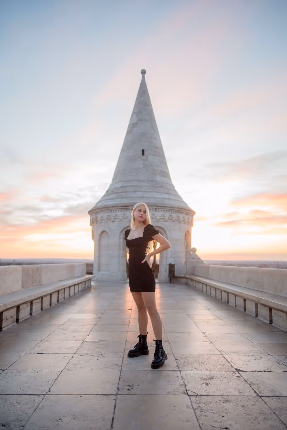 Young woman in a black dress and boots standing on a tiled terrace in front of a white stone tower at sunset.