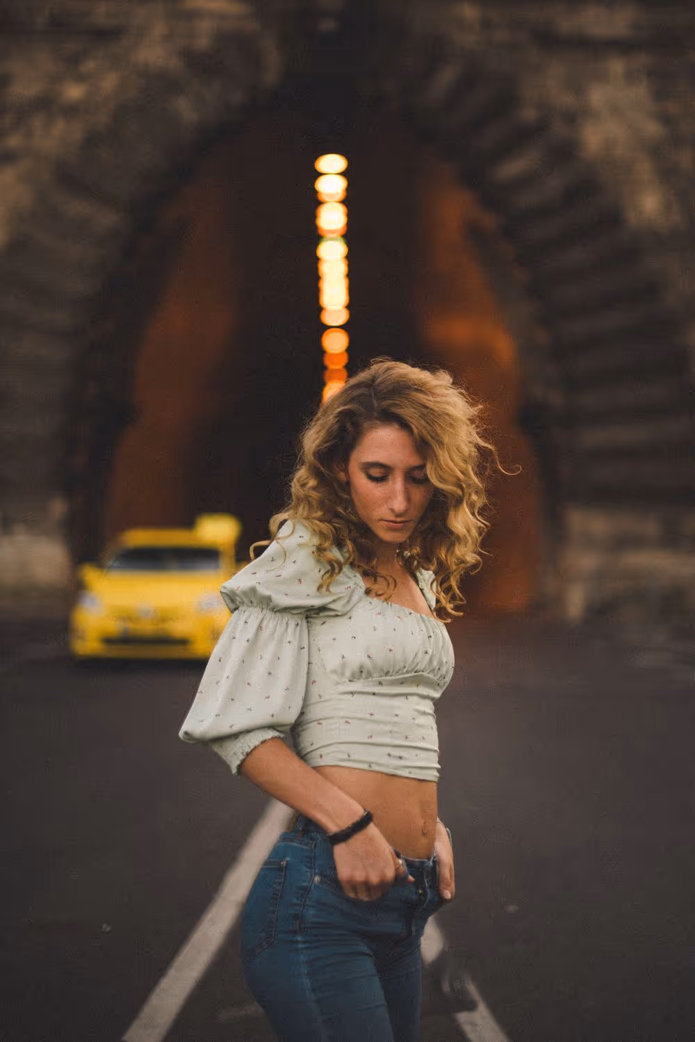 Curly-haired woman in a puffed-sleeve crop top and jeans standing on a road in front of an arched stone tunnel with a yellow car behind her.