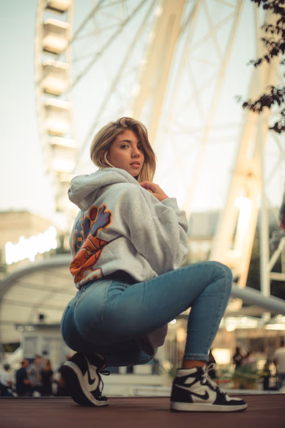 Young woman in a gray hoodie and blue jeans squatting outdoors near a Ferris wheel.