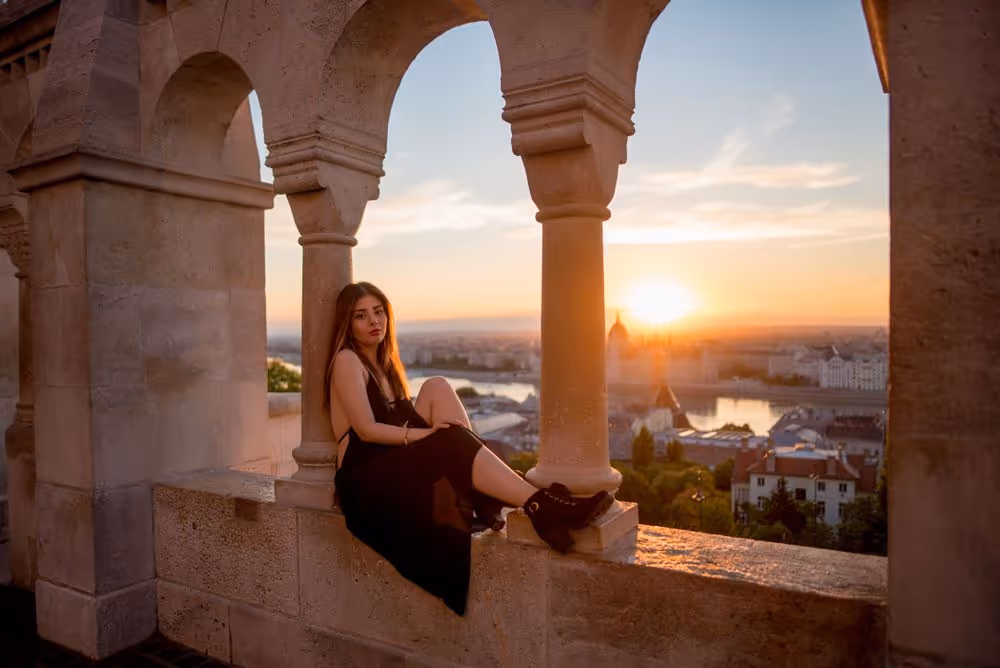 Woman in black dress sitting on stone ledge between arches during sunset overlooking a city with river.