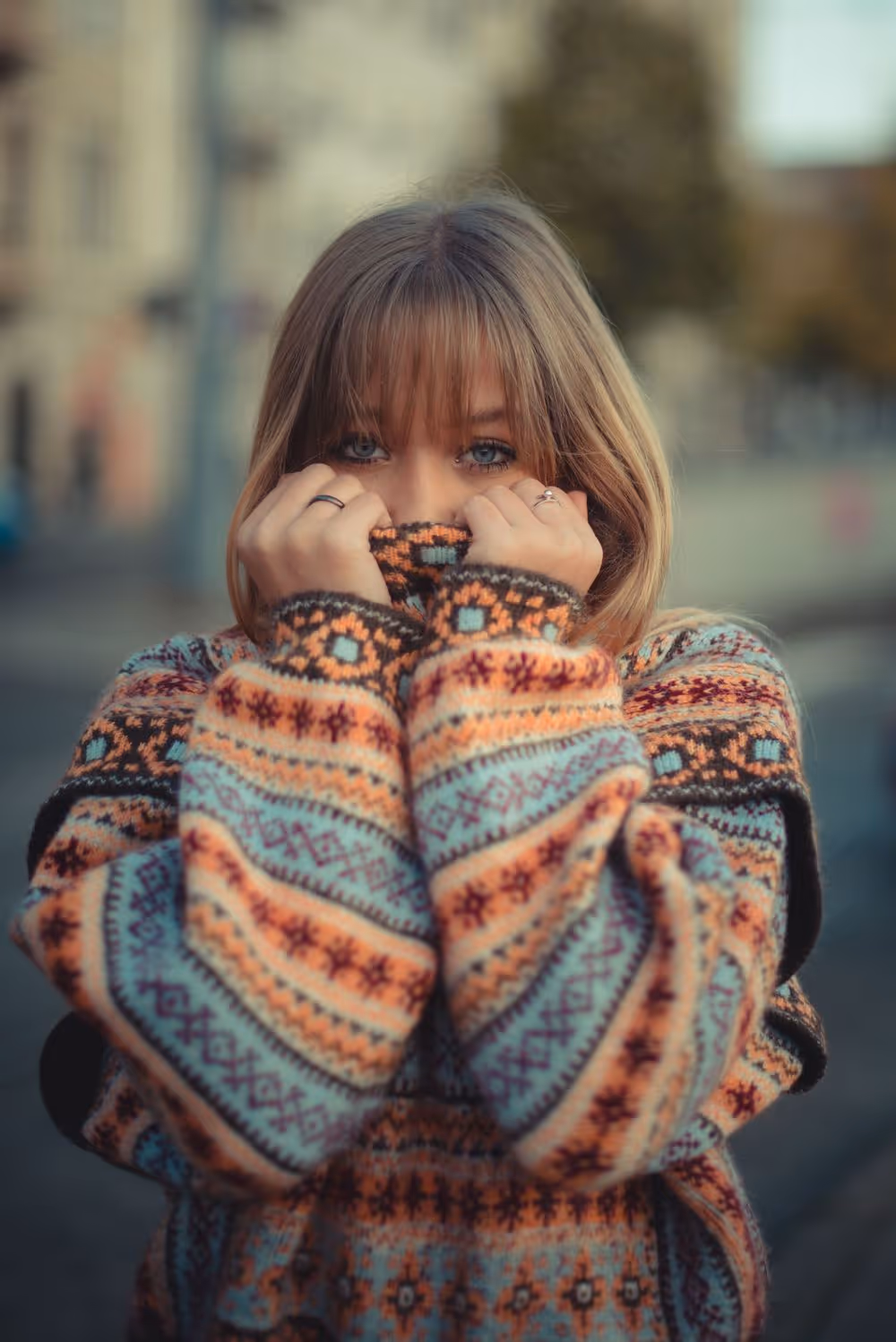 Young woman with blonde hair partially covering her face with the collar of a colorful patterned sweater.