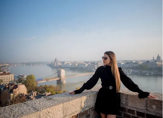 Woman in a black dress with long hair and sunglasses leaning on a stone railing overlooking a river and a city skyline.