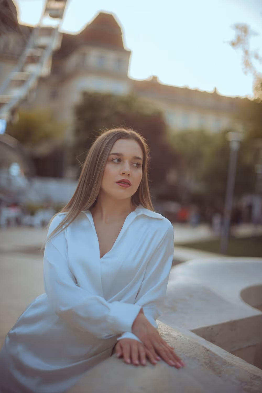 Young woman in a white blouse leaning on a concrete structure outdoors with blurred buildings and trees in the background.