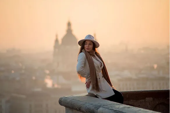 Woman wearing a white hat, beige coat, and brown scarf sitting on a stone ledge with a hazy cityscape and cathedral silhouette in the background at sunset.