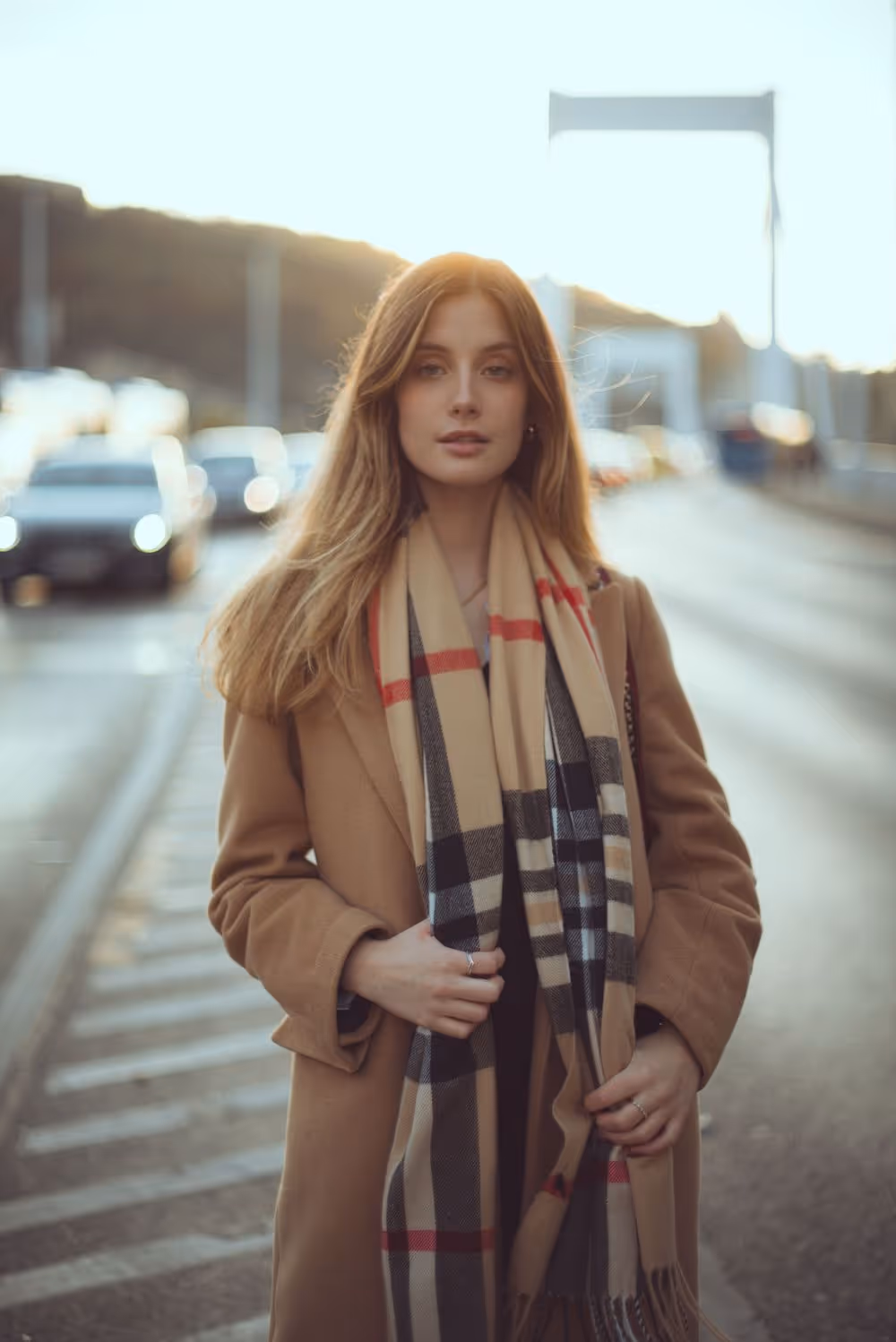 Young woman with long hair wearing a plaid scarf and camel coat standing on a street with blurred cars and bridge in the background at sunset.