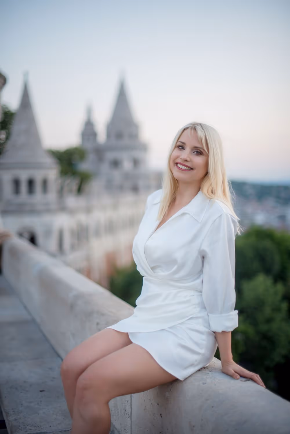 Smiling blonde woman in a white dress sitting on a stone railing with blurred castle towers in the background.
