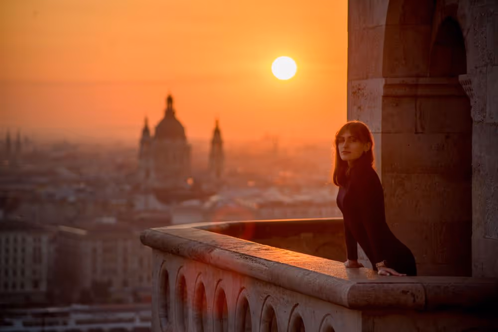 Woman leaning on a stone balcony railing at sunset overlooking a city with a domed building in the background.