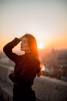 Woman with long hair in dark clothing standing by stone railing during sunset with eyes closed.