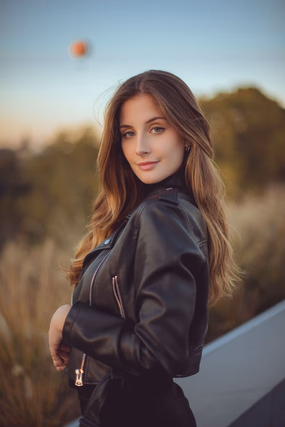 Young woman with long hair wearing a black leather jacket looking over her shoulder outdoors.