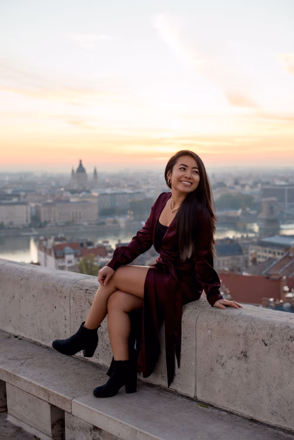 Smiling woman in a dark burgundy dress and black boots sitting on a stone ledge with a cityscape and sunset in the background.