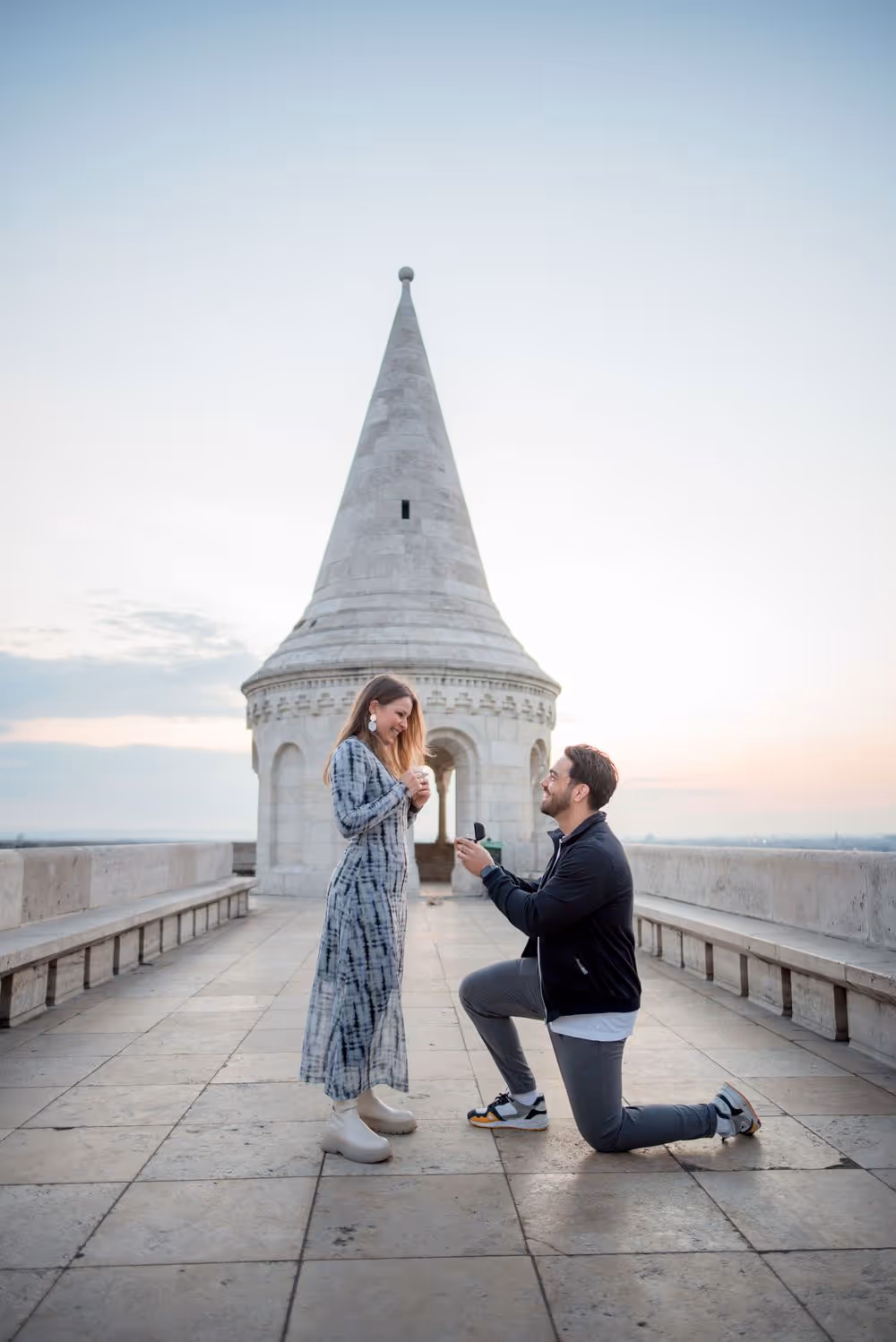 Man kneeling and proposing with an engagement ring to a woman on a stone terrace near a historic tower at sunset.