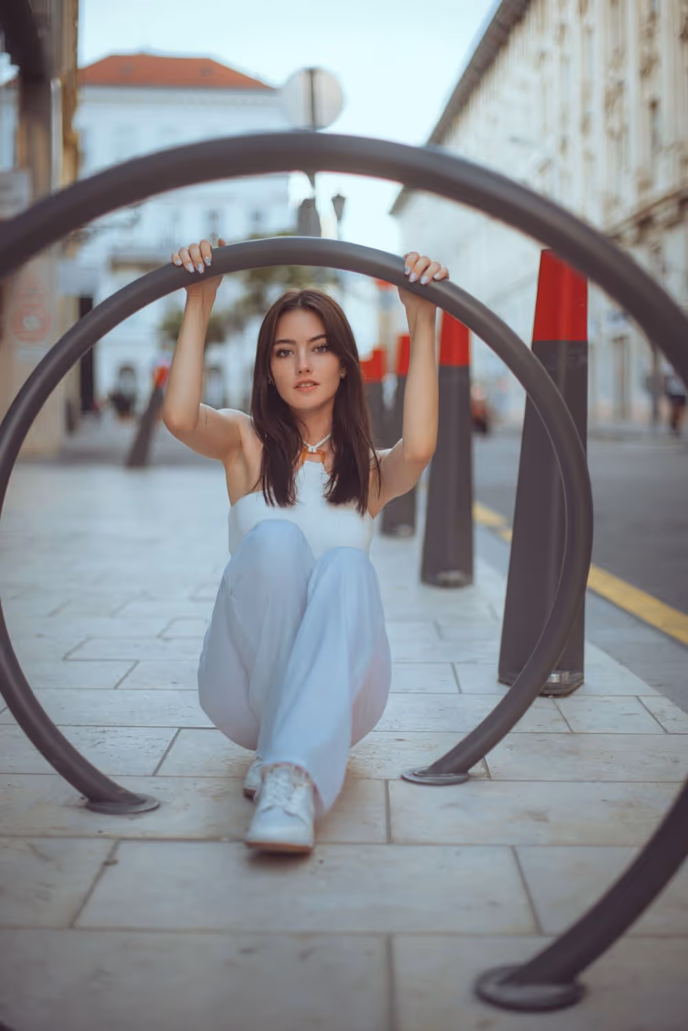 Young woman in white outfit sitting on a city sidewalk holding onto a circular metal bike rack.