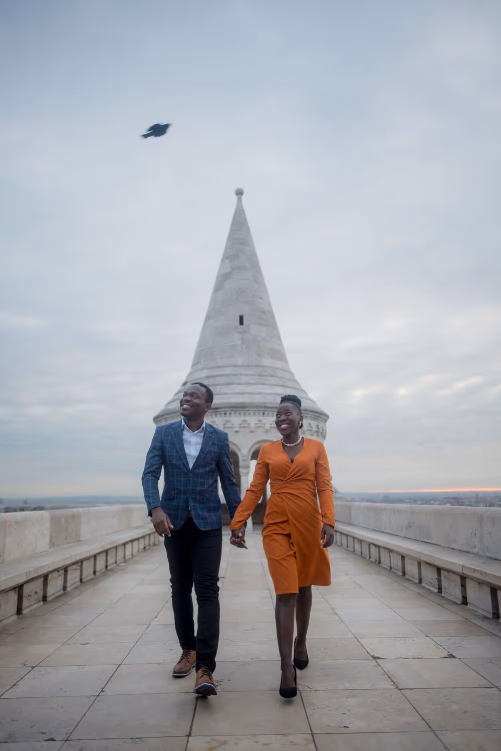 Smiling couple holding hands and walking on a stone pathway with a tall white tower and cloudy sky in the background.