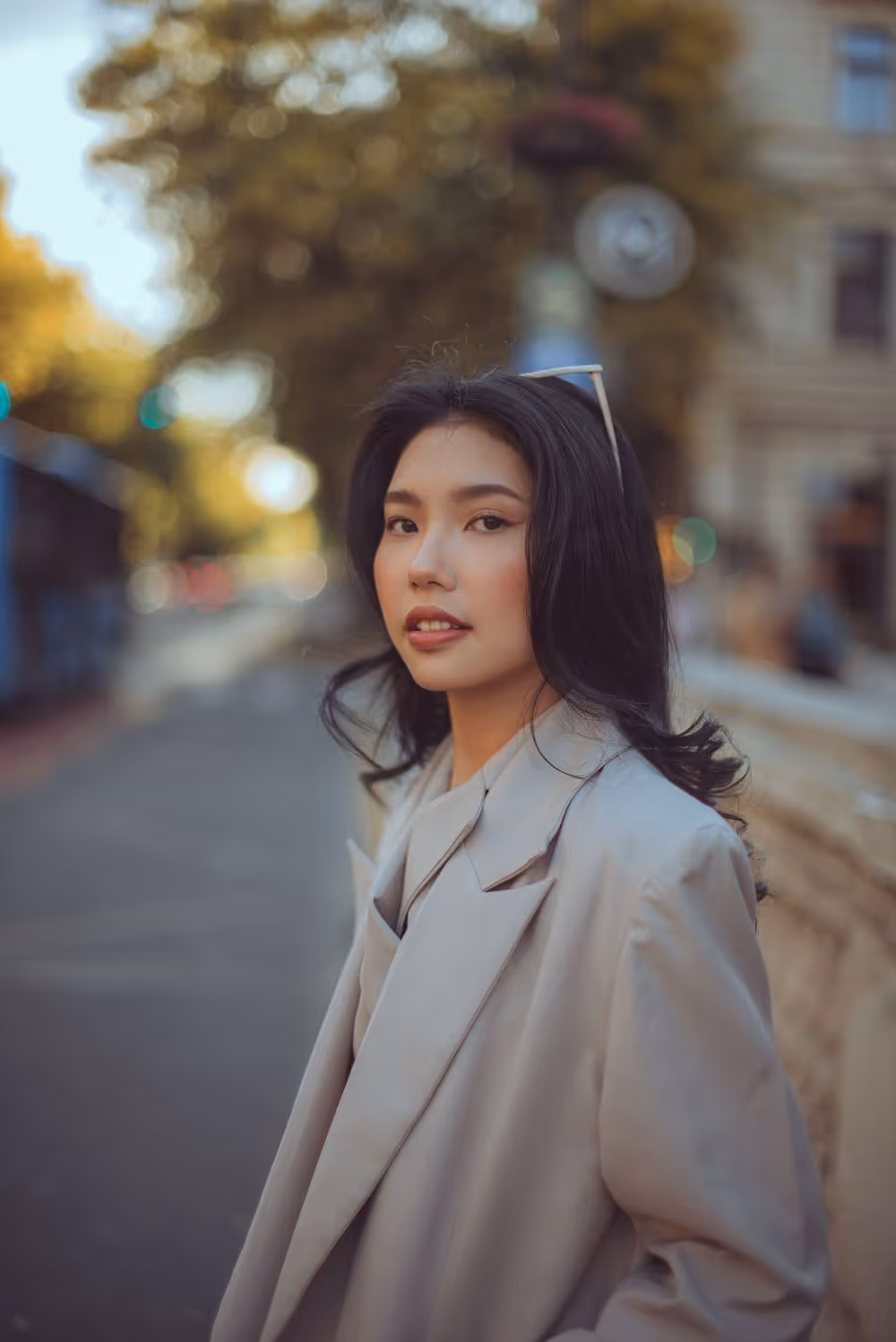 Young woman with long black hair wearing a light gray coat and sunglasses on her head, standing on a city street with blurred trees and buildings in the background.