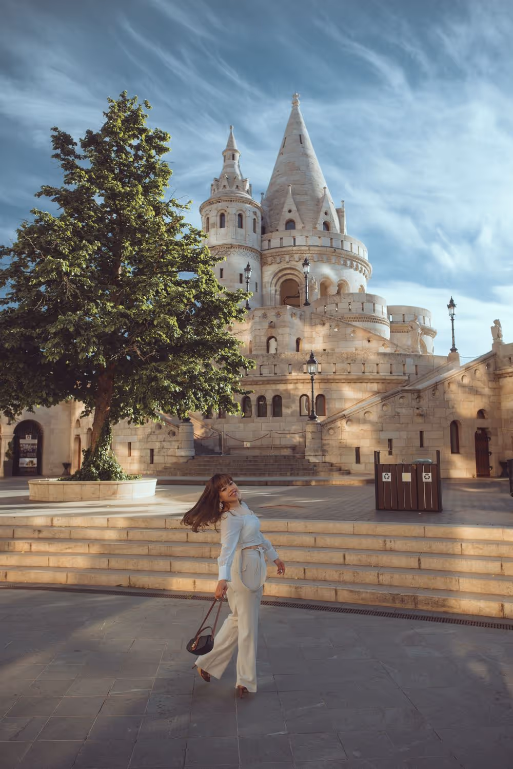 Smiling woman in light outfit walking near a large tree and historic stone castle with towers under a blue sky.