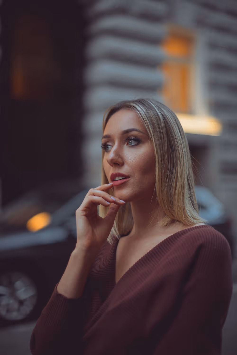Blonde woman in a brown sweater looking thoughtfully to the side with her finger near her lips against a blurred urban background.
