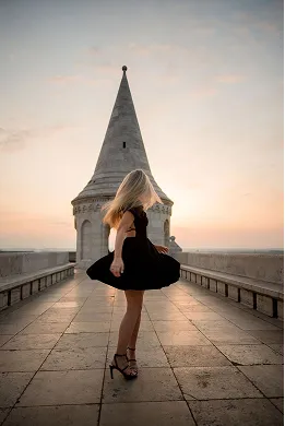 Woman in a black dress twirling on a stone walkway in front of a white tower at sunset.