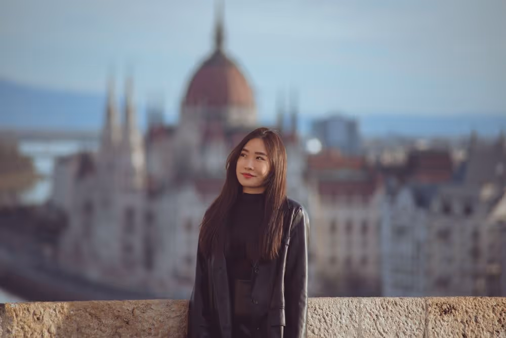Young woman with long dark hair and black jacket standing outdoors with blurred historic building in the background.
