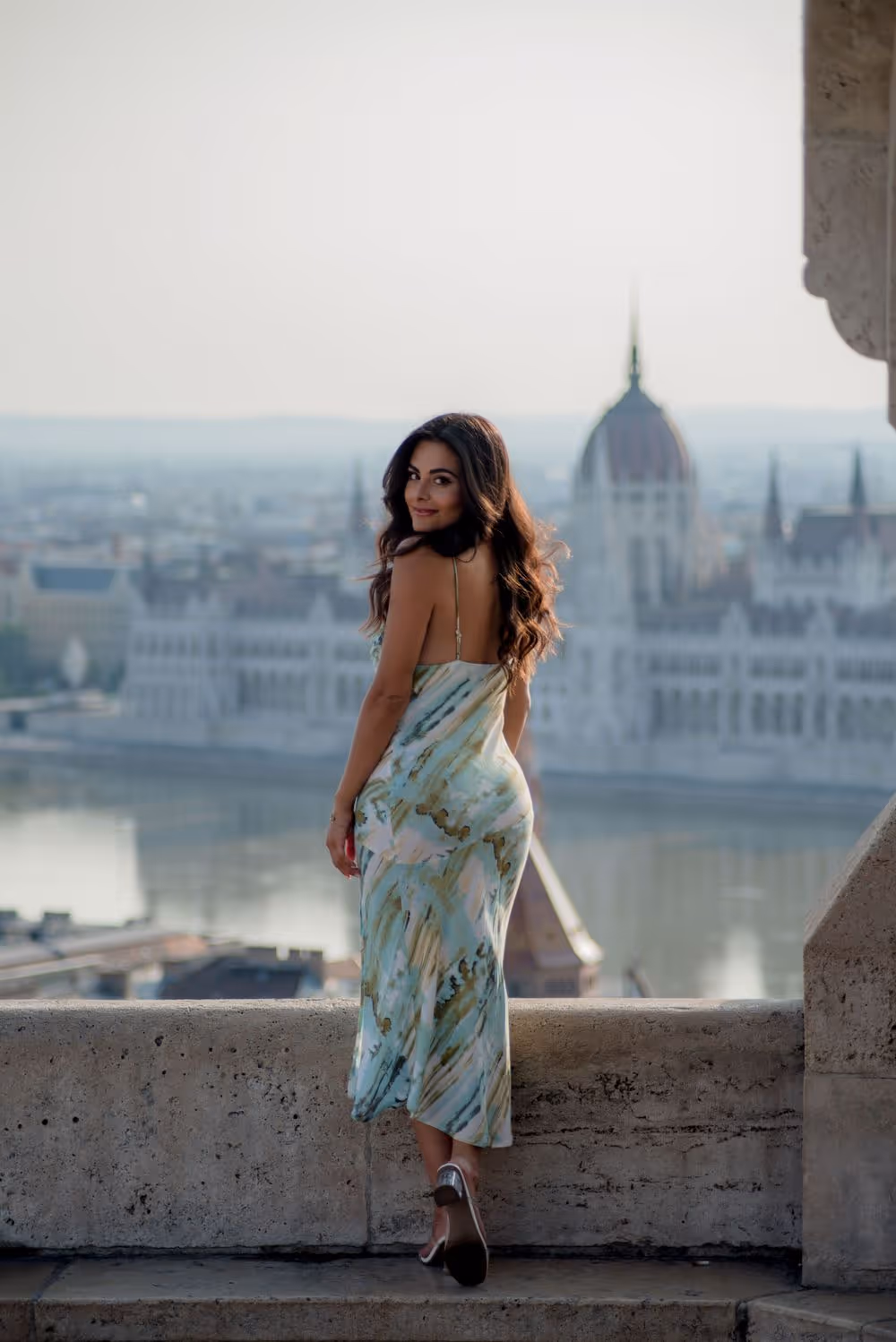 Woman in a light blue and green dress standing on a stone balcony overlooking a river and historic buildings.