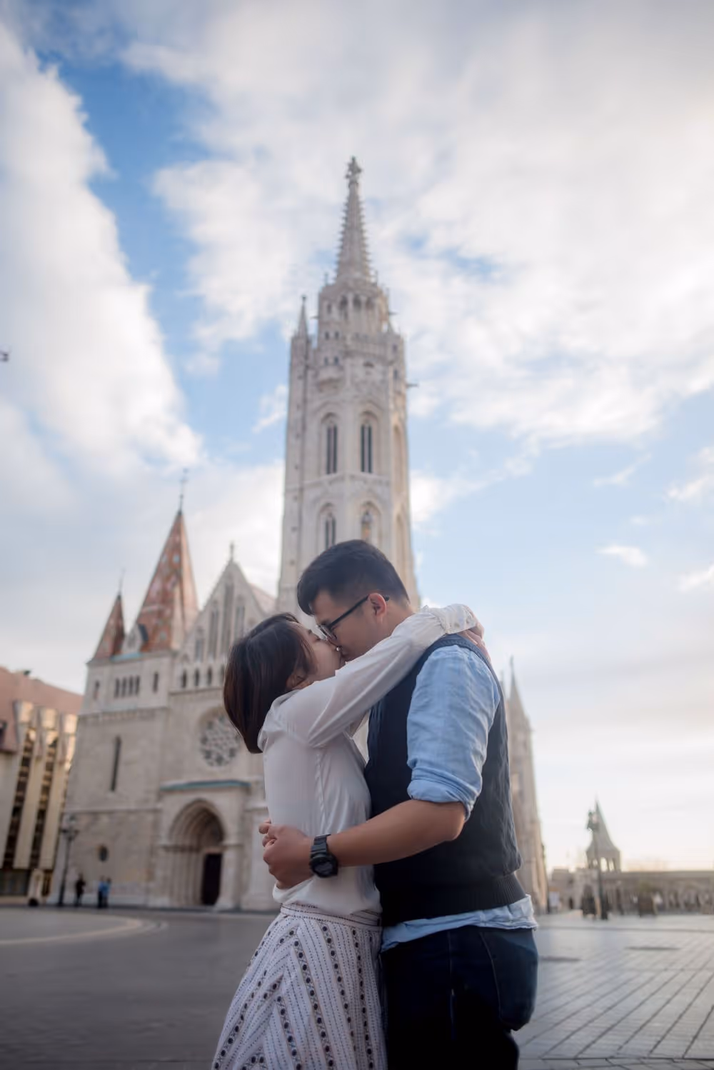 Couple embracing and kissing in front of a historic Gothic-style church with a tall spire under a partly cloudy sky.