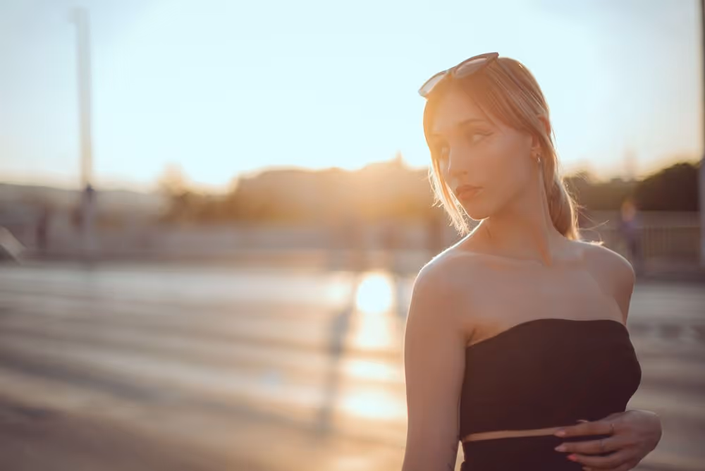 Young woman with sunglasses on her head wearing a strapless black top, standing outdoors with warm sunlight in the background.