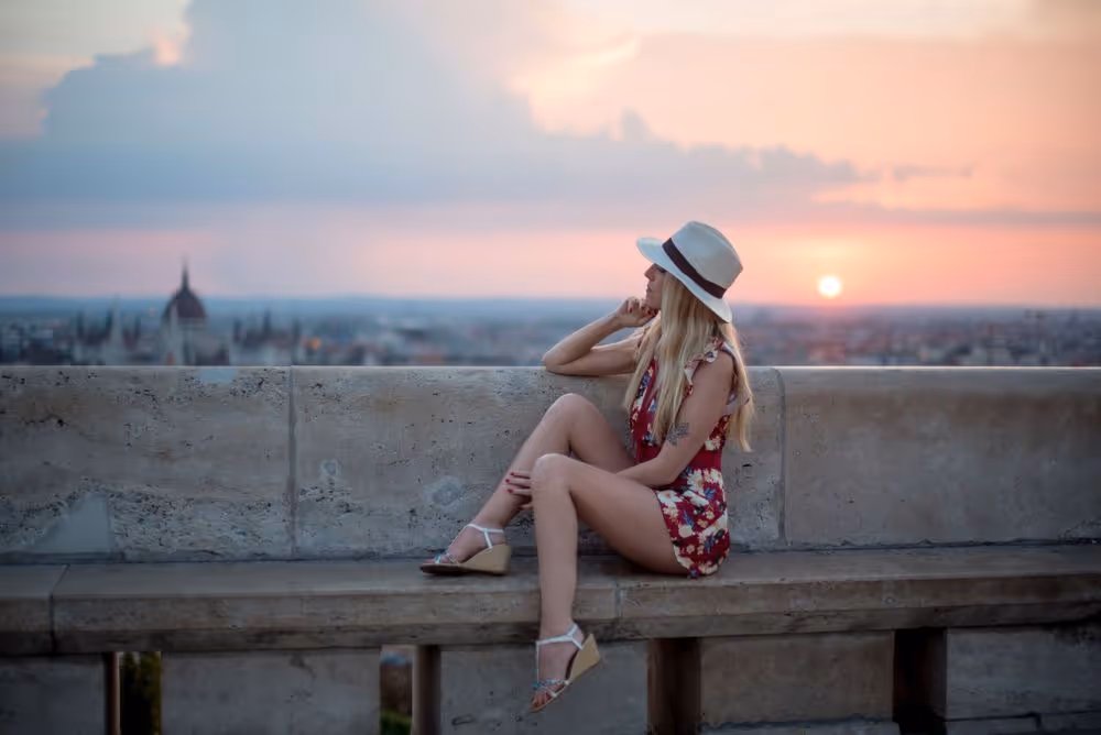 Woman in a floral dress and hat sitting on a stone bench at sunset, overlooking a cityscape.