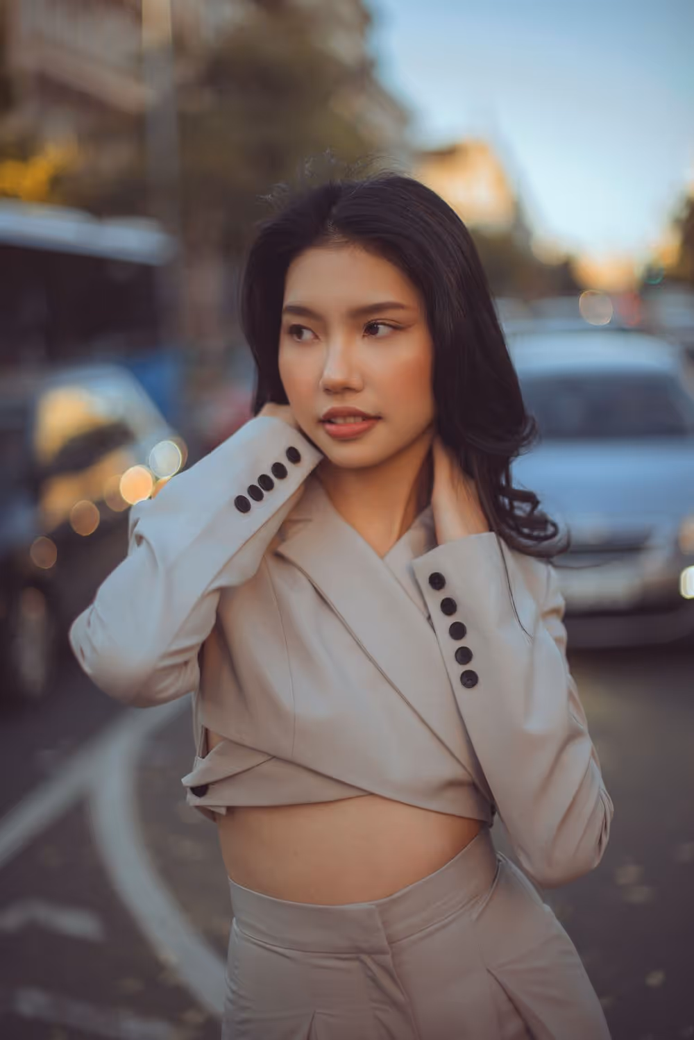 Young woman with long dark hair wearing a beige cropped blazer and matching pants, posing outdoors on a city street with blurred cars in the background.