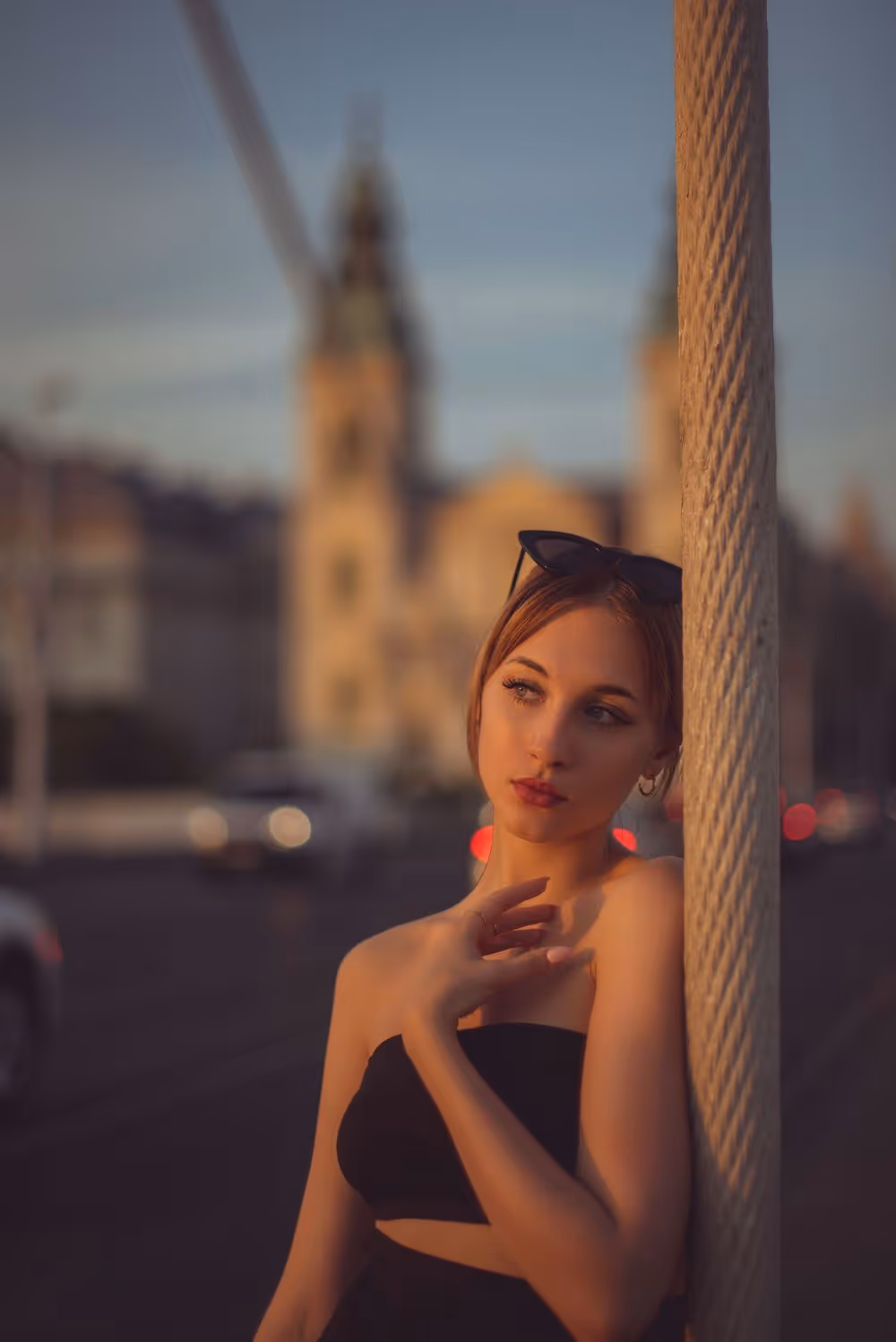 Young woman in black strapless top leaning against a textured pole with a blurred cityscape background at sunset.