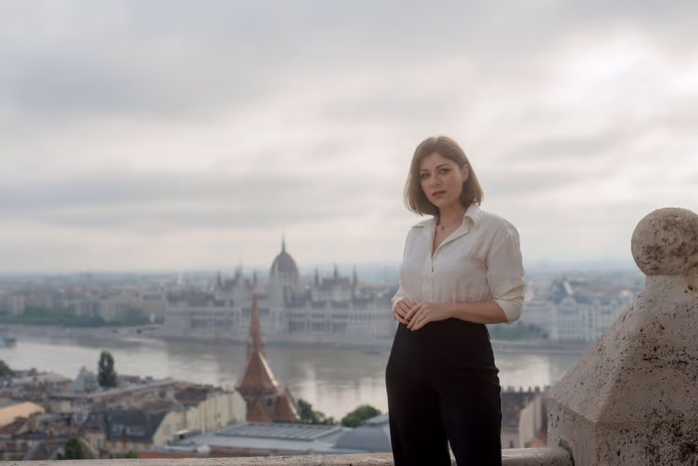Woman in white blouse and black pants standing on a balcony overlooking a river and historic cityscape with a cloudy sky.