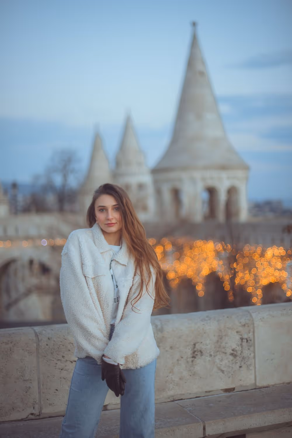 Young woman in a white fuzzy jacket and gloves standing in front of stone railing with blurred historic towers and warm string lights in the background.