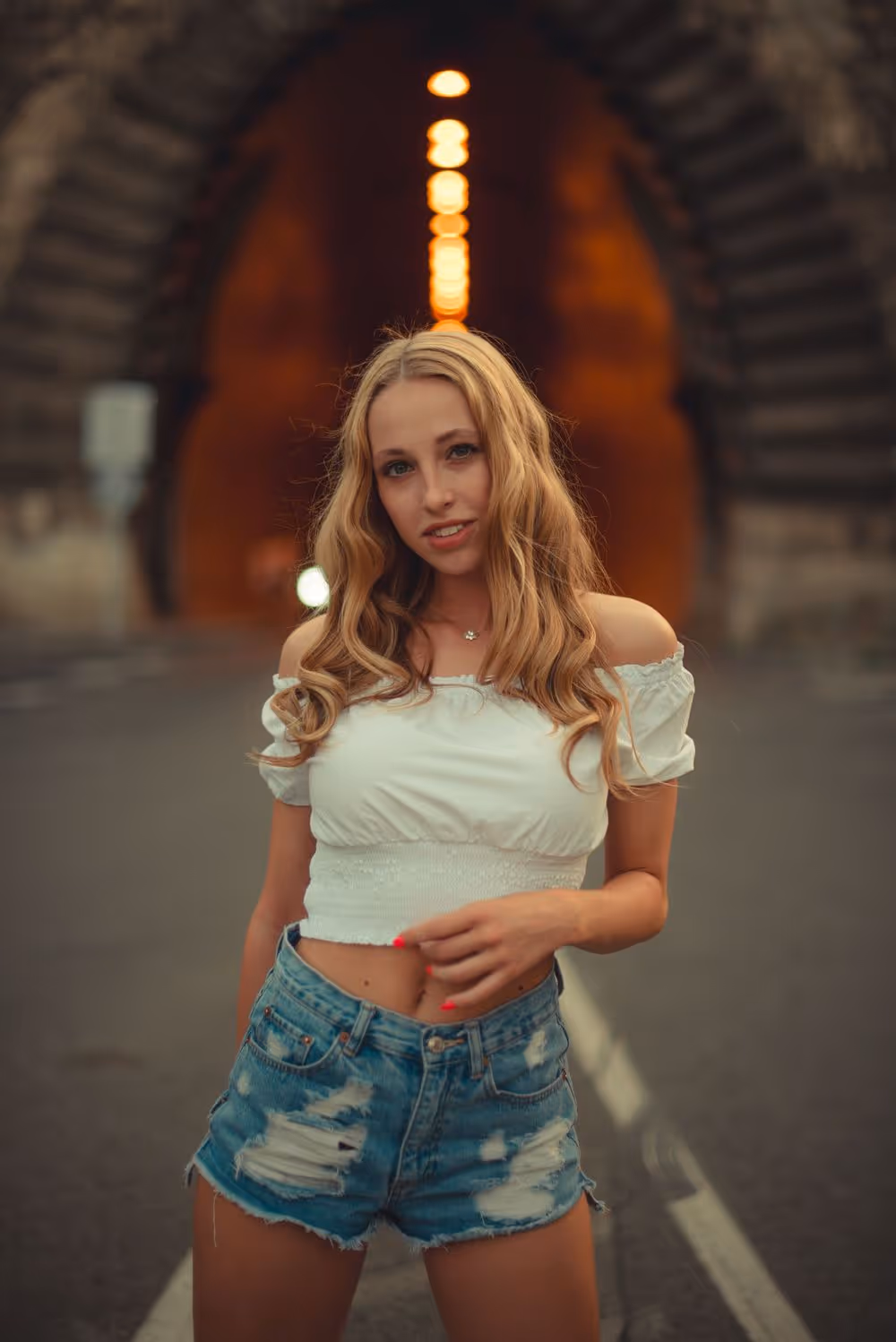 Young woman with long blonde hair wearing a white off-shoulder top and ripped denim shorts standing on a road in front of an illuminated stone tunnel.