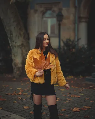 Woman wearing a yellow jacket and black dress holding a large autumn leaf outdoors in a park.