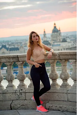 Woman in beige crop top and black pants leaning on stone railing with cityscape and domed building at sunset in the background.