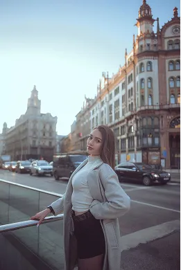 Young woman in a light coat posing by a glass railing on a city street with historic buildings and cars in the background during daylight.
