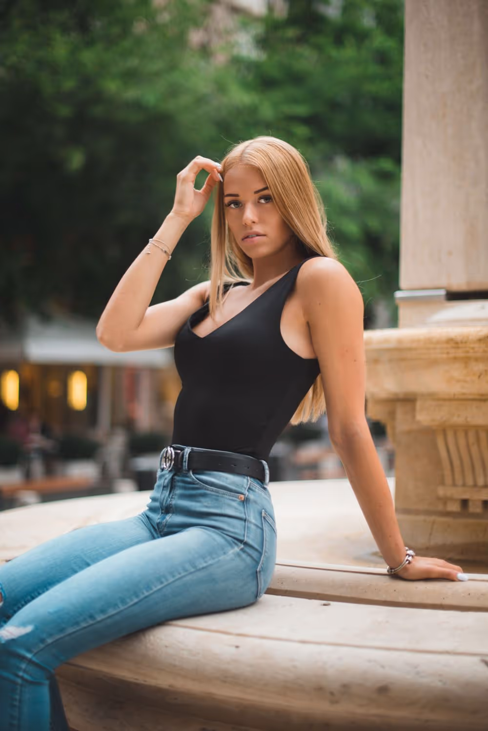 Young woman with long blonde hair in a black sleeveless top and blue jeans sitting on a stone ledge outdoors.