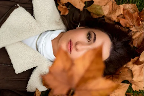 Woman lying on ground covered with autumn leaves, holding a large brown leaf in front of her face.